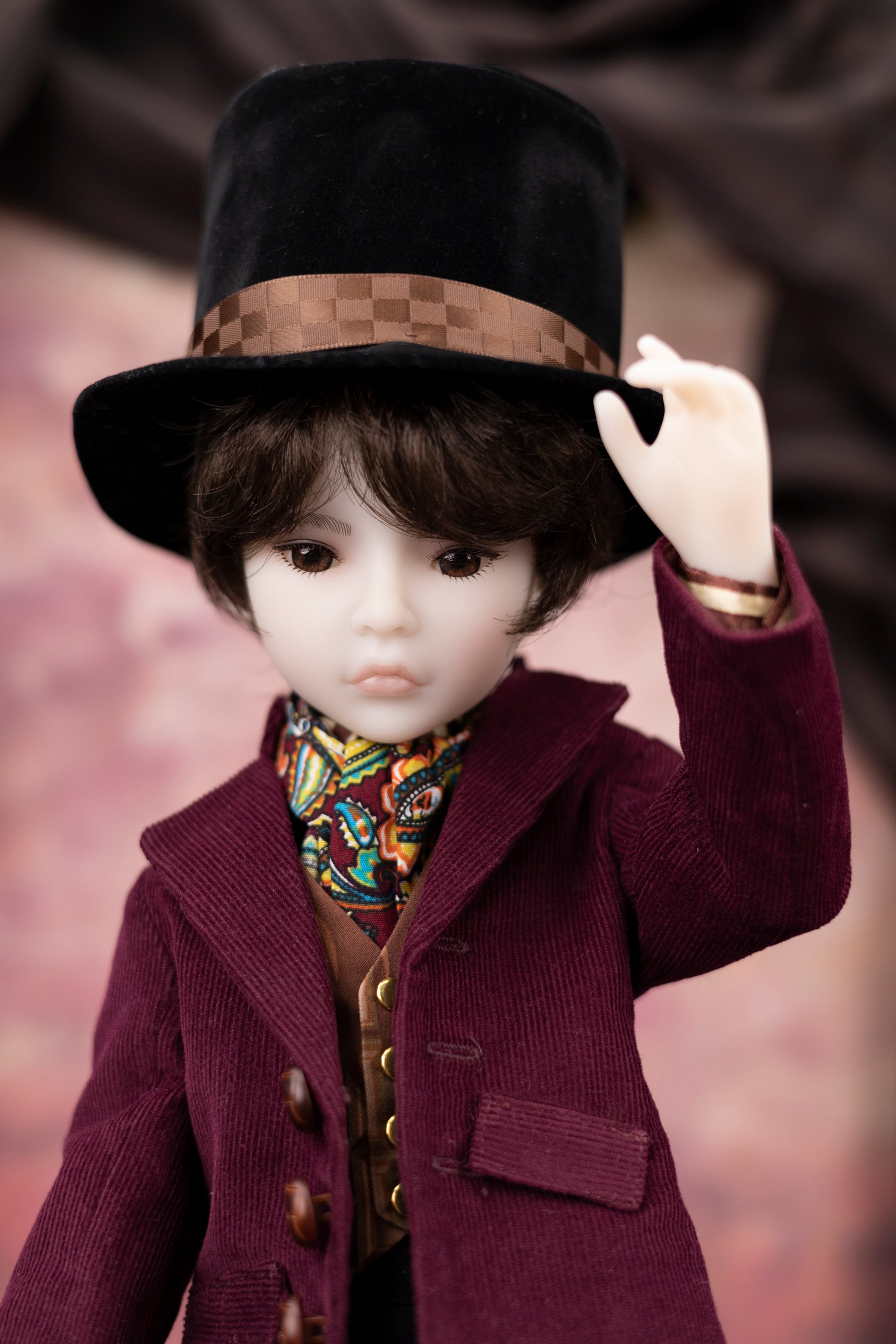 Doll wearing a black hat and  burgundy red corduroy coat with a blurred background