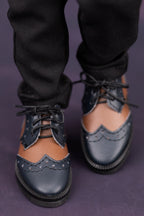 Black leather doll shoes with brogue details worn with black pants on a dark background