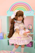 Doll in a pastel dress holding a cake in front of a rainbow-colored 'Shop' sign.