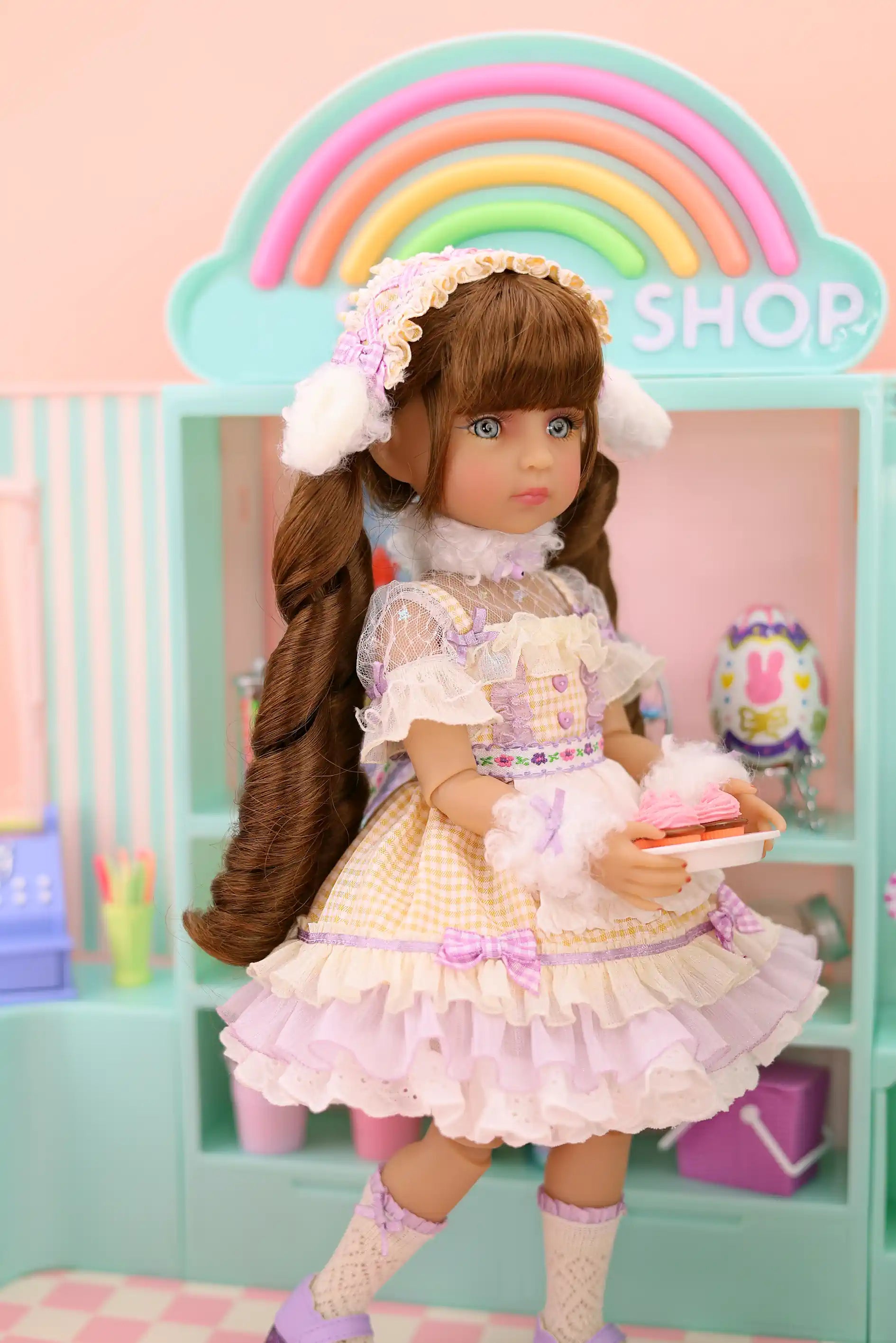 Doll in a pastel dress holding a cake in front of a rainbow-colored 'Shop' sign.