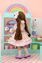 Doll with long brown hair in a white dress standing in front of a colorful playset with a rainbow.