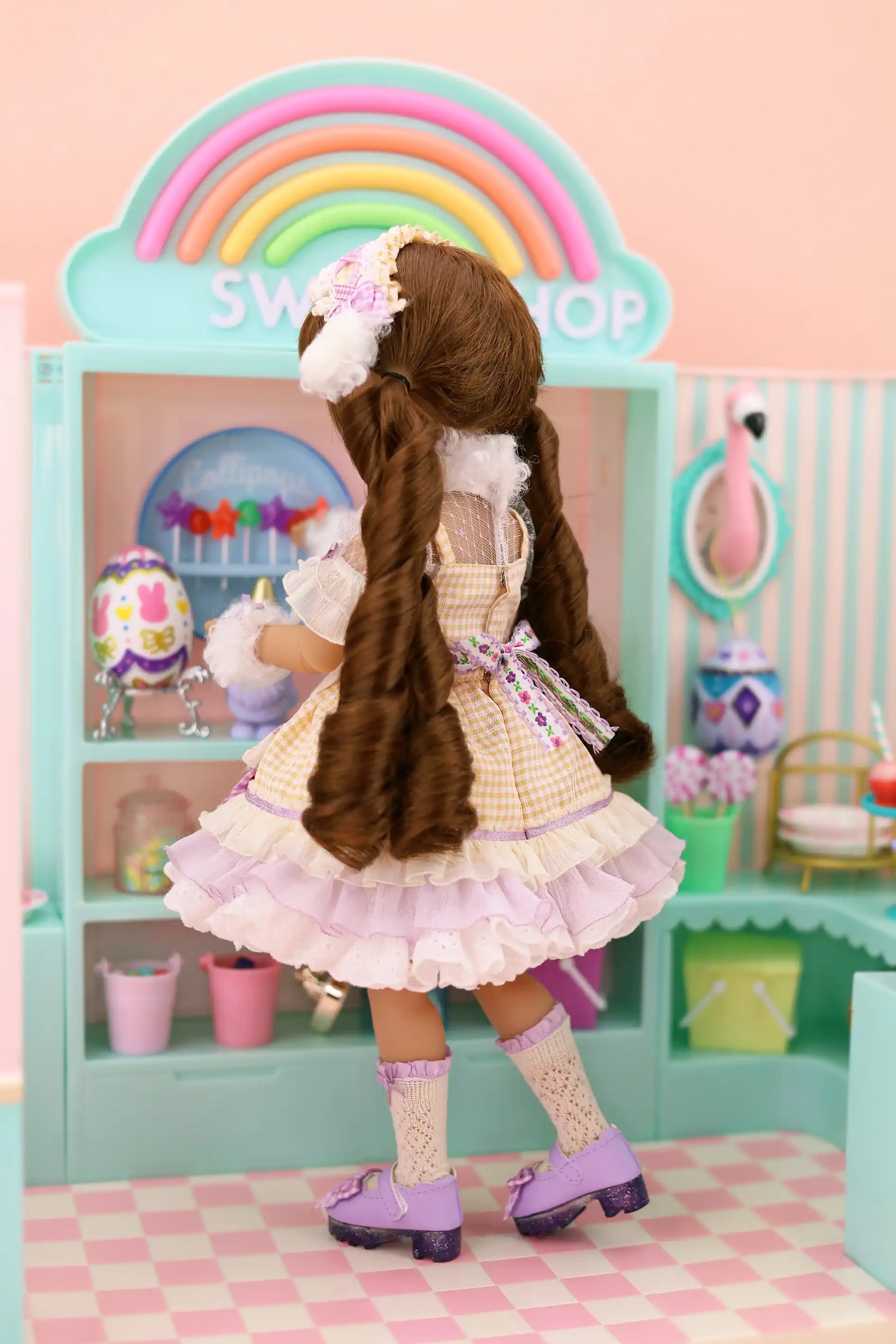 Doll with long brown hair in a white dress standing in front of a colorful playset with a rainbow.
