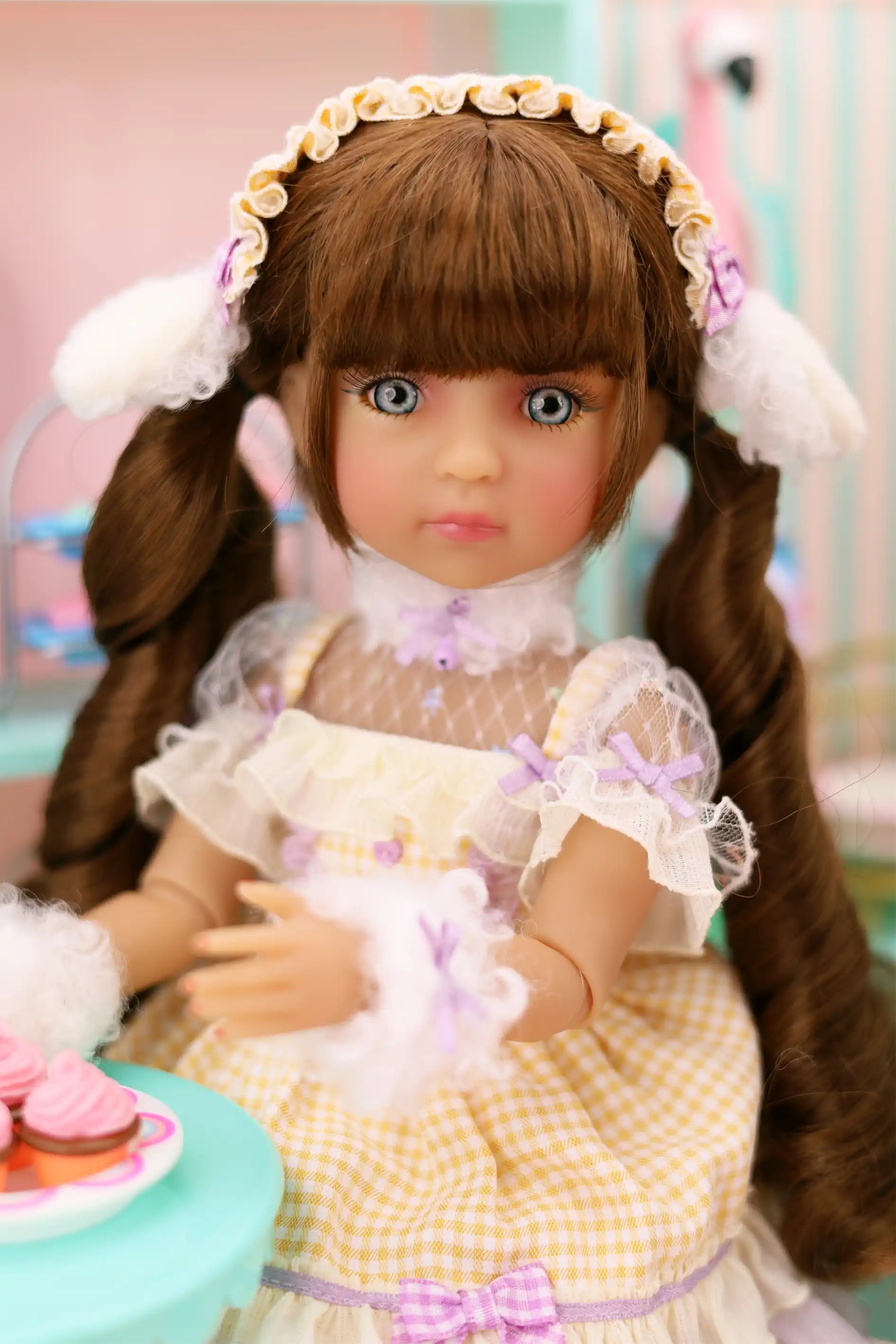 Doll with long brown hair styled in pigtails, wearing a yellow dress with white lace and a headband with white flowers.