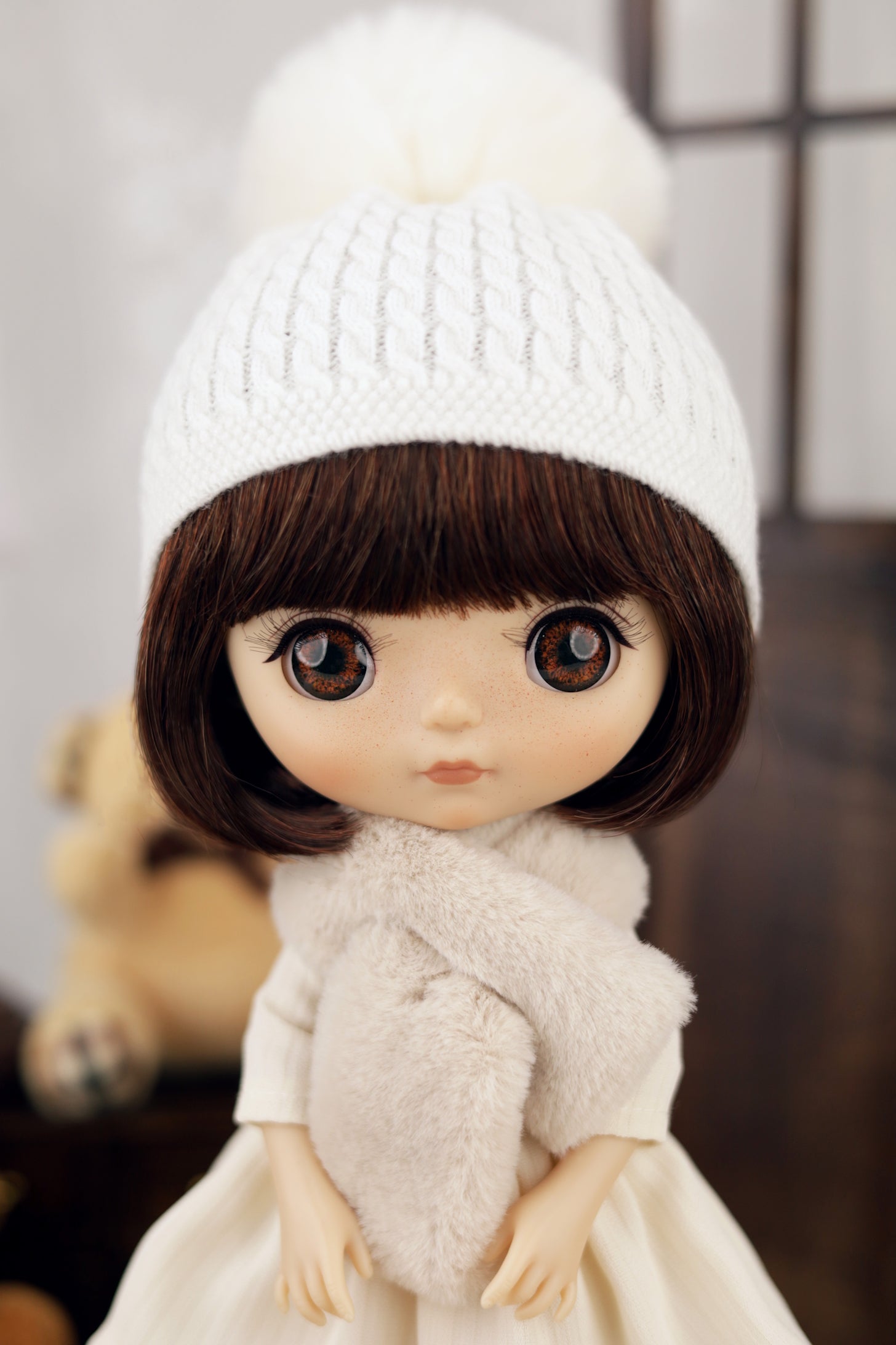 Doll wearing a white knit hat and scarf with a blurred background
