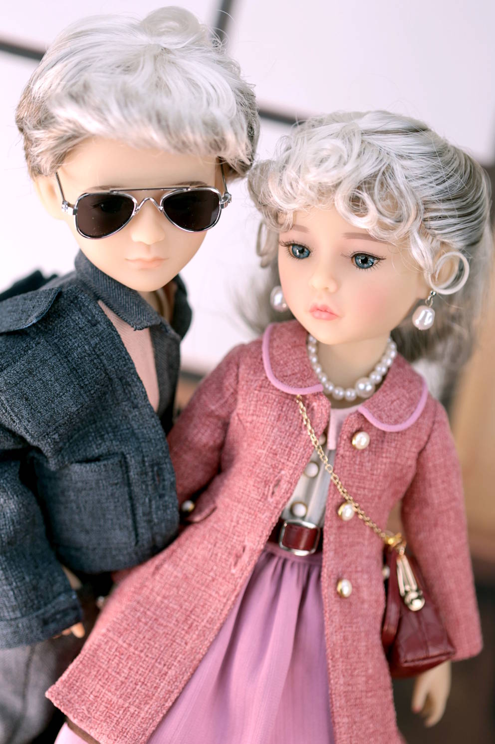 Two dolls, one with sunglasses and a gray coat, the other in a pink dress with a pearl necklace.