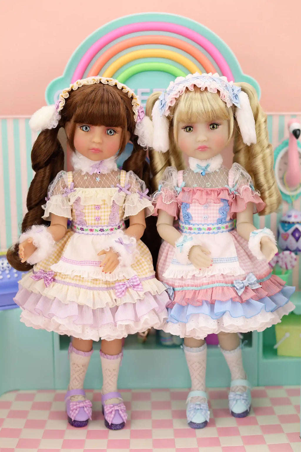 Two dolls in pastel outfits standing in front of a rainbow, with a pink checkered floor and light blue wall in the background.