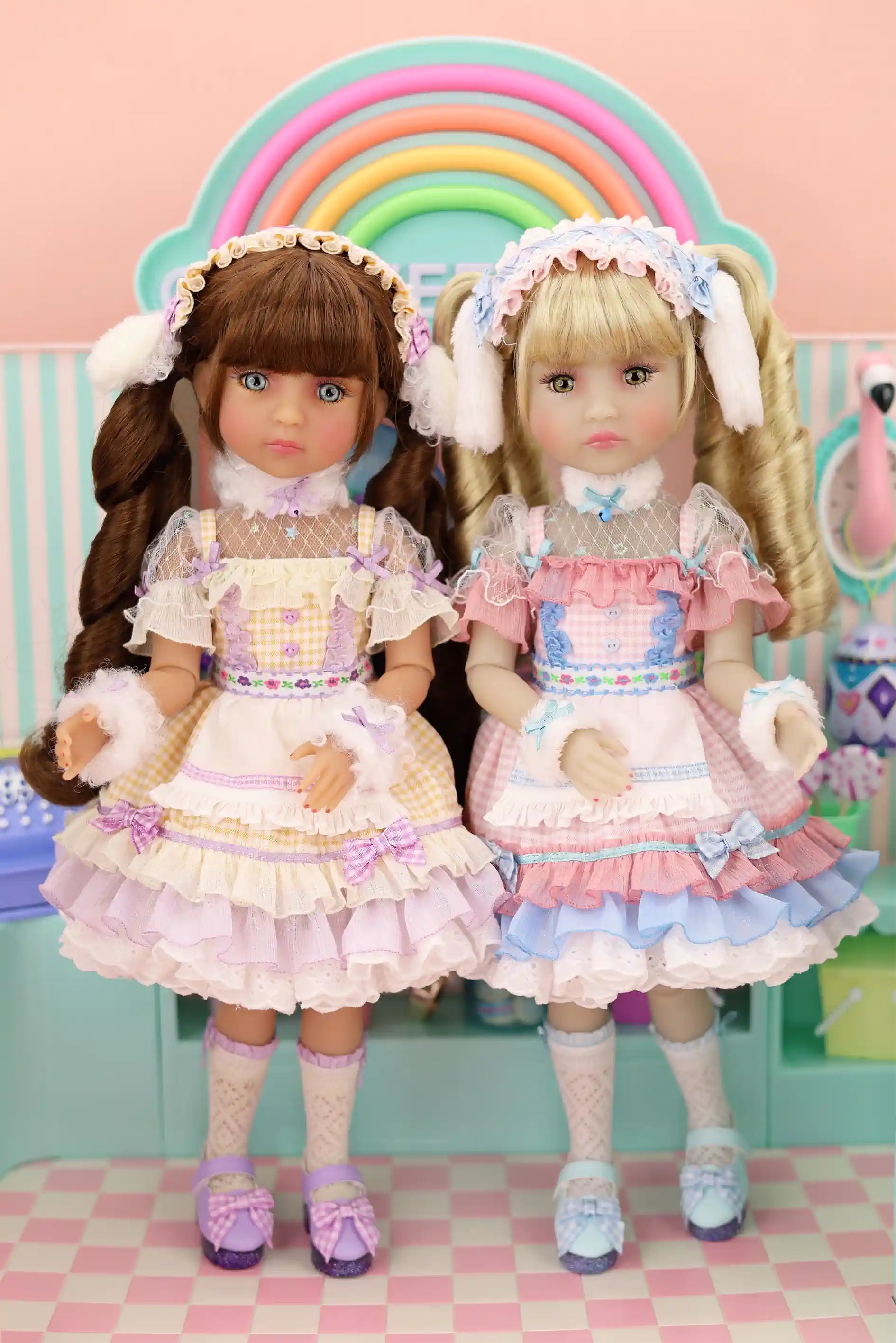 Two dolls in pastel outfits standing in front of a rainbow, with a pink checkered floor and light blue wall in the background.